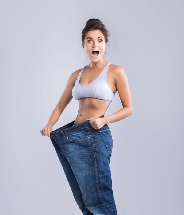 Eine Frau in einem Sport-BH hält eine übergroße Jeans in die Höhe und wirkt überrascht, was einen deutlichen Gewichtsverlust vor einem schlichten Hintergrund verdeutlicht. grüner tee zum abnehmen.