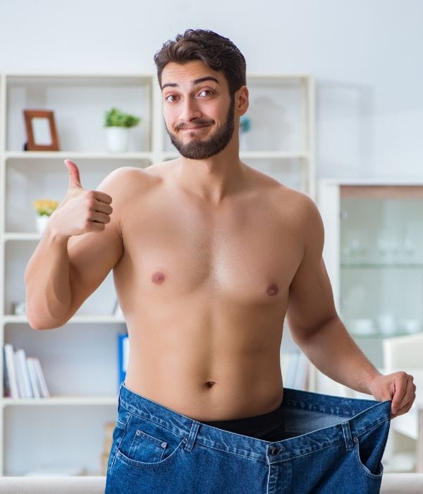 Ein Mann ohne Hemd, der den Daumen nach oben streckt und eine übergroße Jeans trägt, was auf einen erheblichen Gewichtsverlust schließen lässt, steht in einem Raum mit Regalen im Hintergrund. grüner tee zum abnehmen.