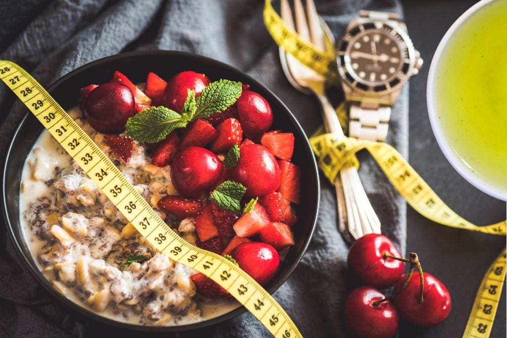 Eine Schale Chia-Pudding mit Kirschen, Erdbeeren und Minze, daneben ein gelbes Maßband, eine Uhr, Gabeln und ein Becher mit grünem Tee. grüner tee zum abnehmen.