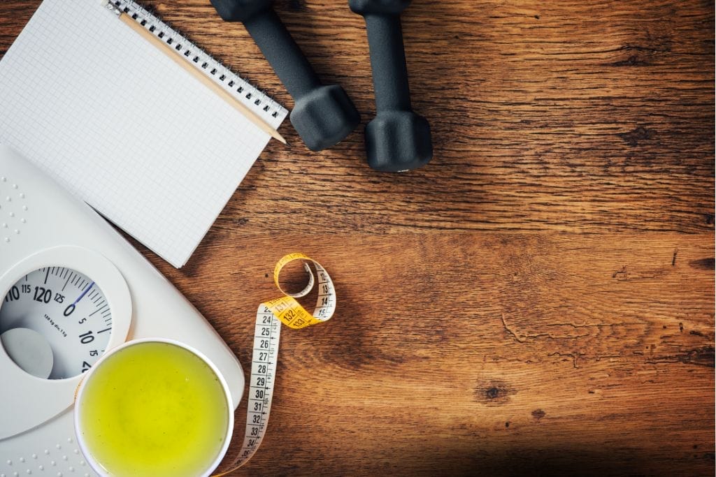 Eine Waage, ein Becher mit grünem Tee, ein Maßband, ein Notizblock und zwei Hanteln sind auf einer Holzfläche angeordnet. grüner tee zum abnehmen.