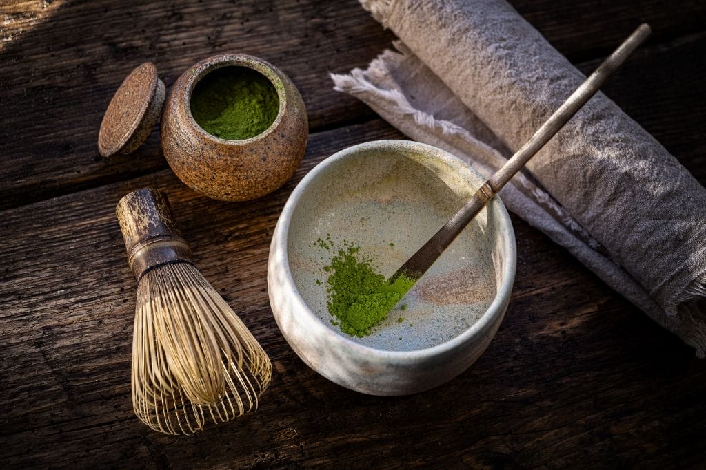 Eine Keramikschale mit Matcha-Pulver und ein Bambusbesen stehen auf einer hölzernen Unterlage, daneben ein kleines Glas mit Matcha-Pulver und eine gerollte Stoffserviette. grüner tee zum abnehmen