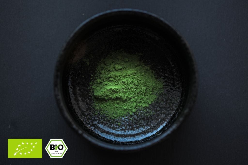 Eine schwarze Schale mit grünem Matcha-Pulver auf einer dunklen Oberfläche. In der unteren linken Ecke sind zwei Logos für die Bio-Zertifizierung abgebildet.